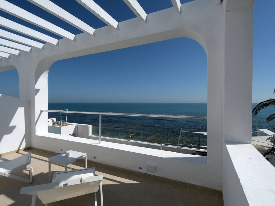 A balcony with white furniture, a clear view of the sea, and a palm tree.
