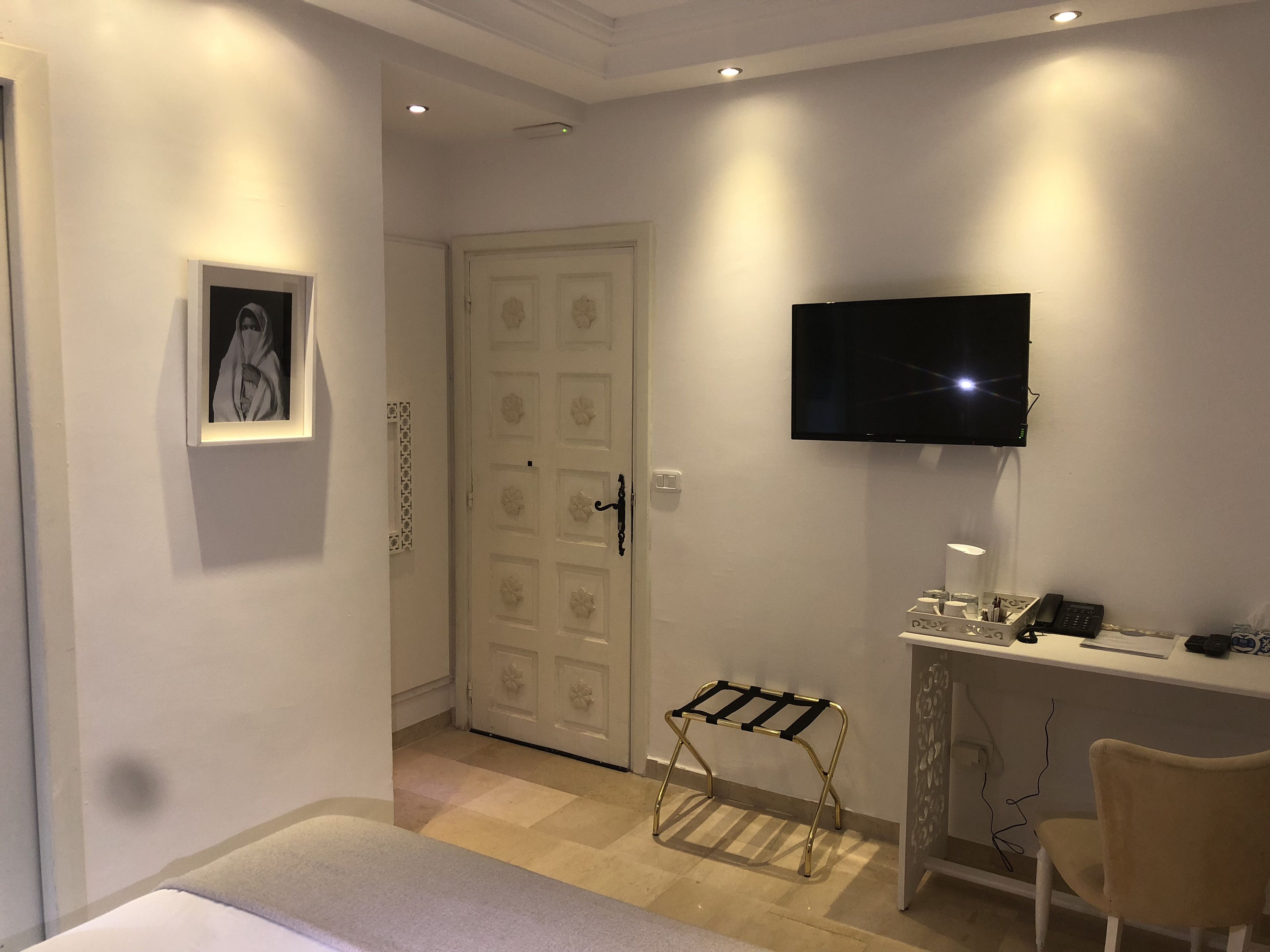 A hotel room with a bed, a desk with a computer, a TV mounted on the wall, a chair, and a door with a decorative pattern.