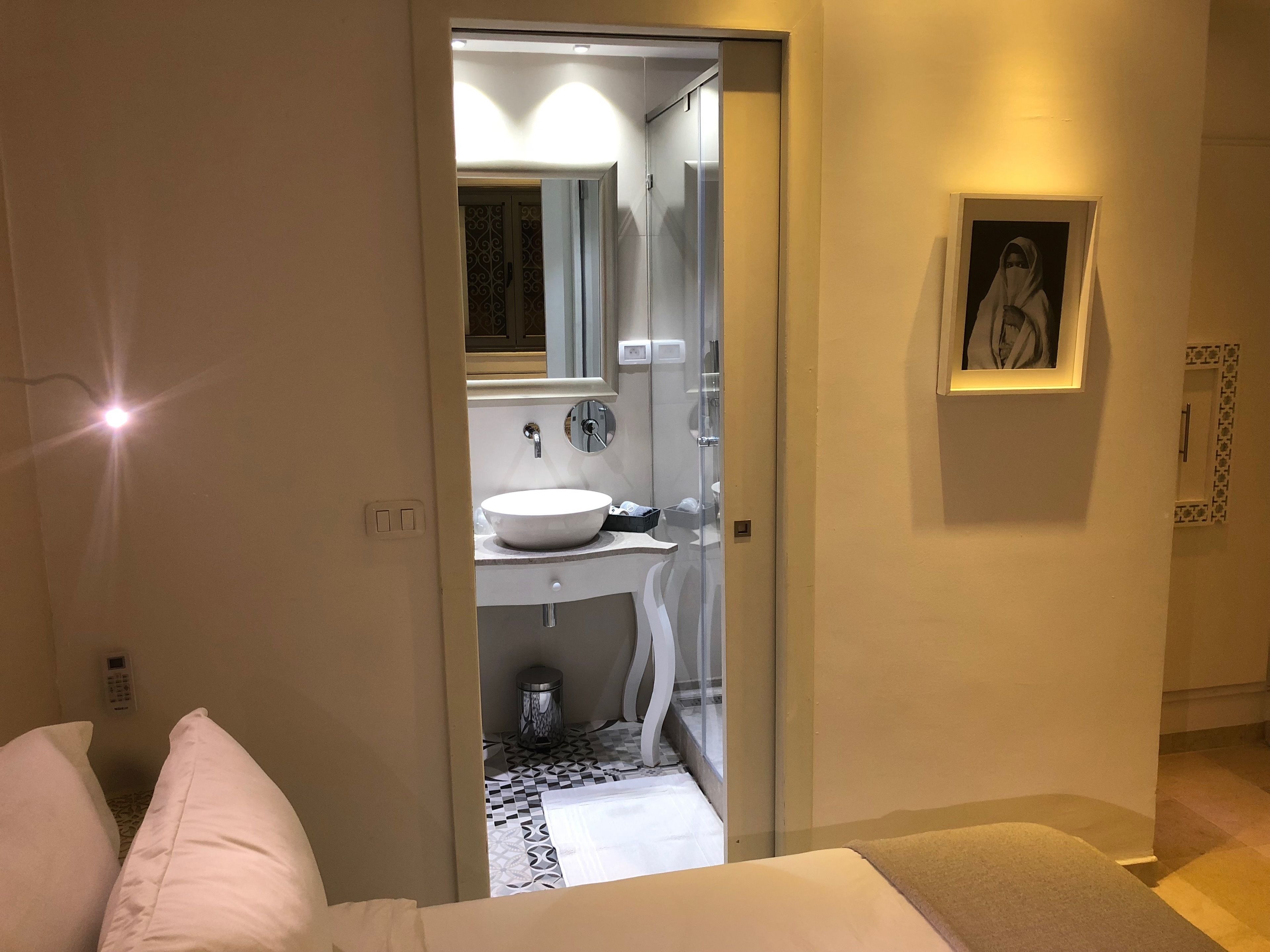 Un baño moderno con un lavamanos blanco, espejo y un cubo de basura pequeño. Hay un cuadro en la pared y una cama con ropa de cama blanca al frente.