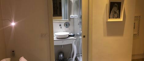 Un baño moderno con un lavamanos blanco, espejo y un cubo de basura pequeño. Hay un cuadro en la pared y una cama con ropa de cama blanca al frente.