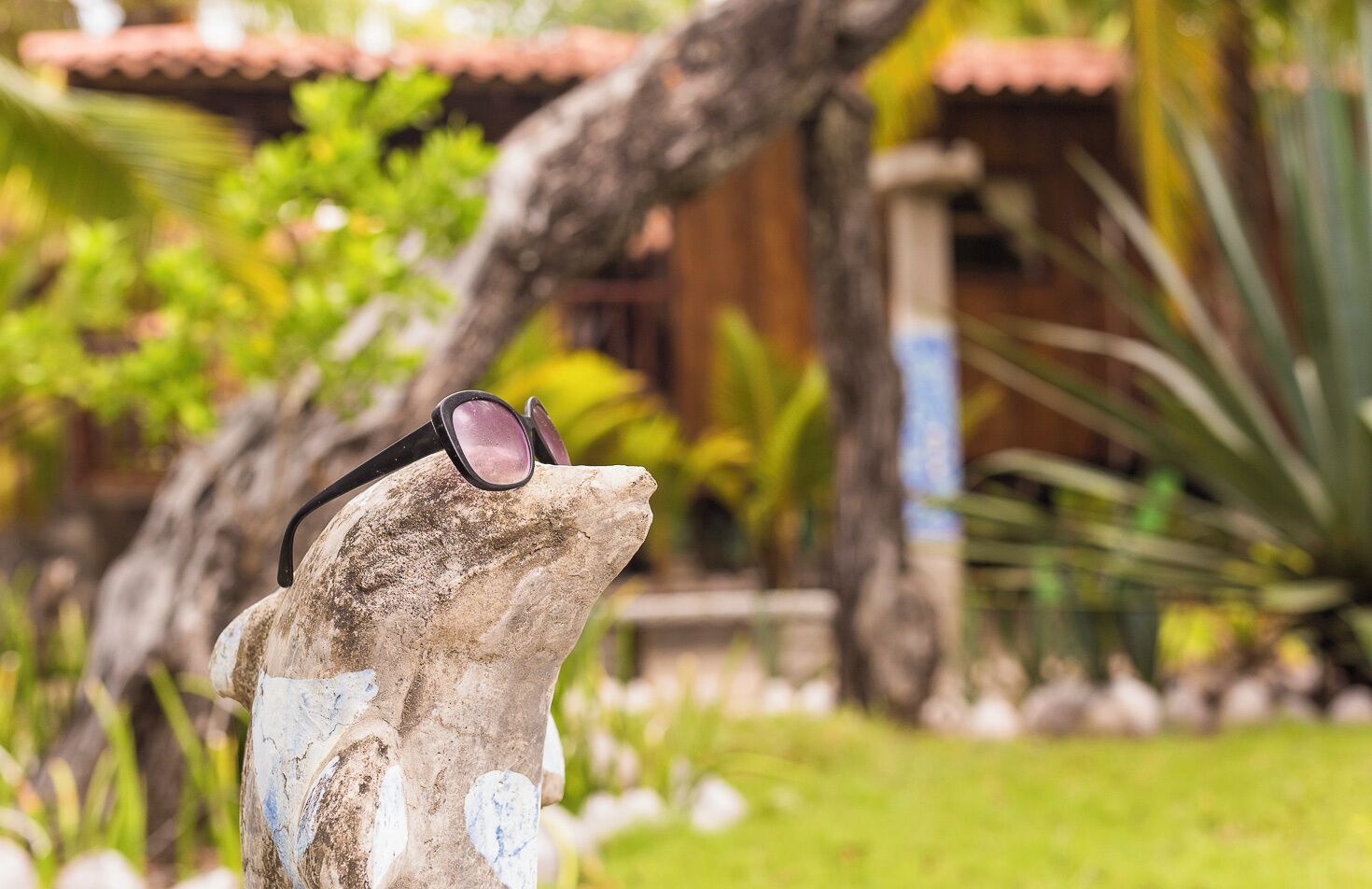 Une statue de pierre portant des lunettes de soleil, placée devant un jardin avec un bâtiment et des arbres en arrière-plan.