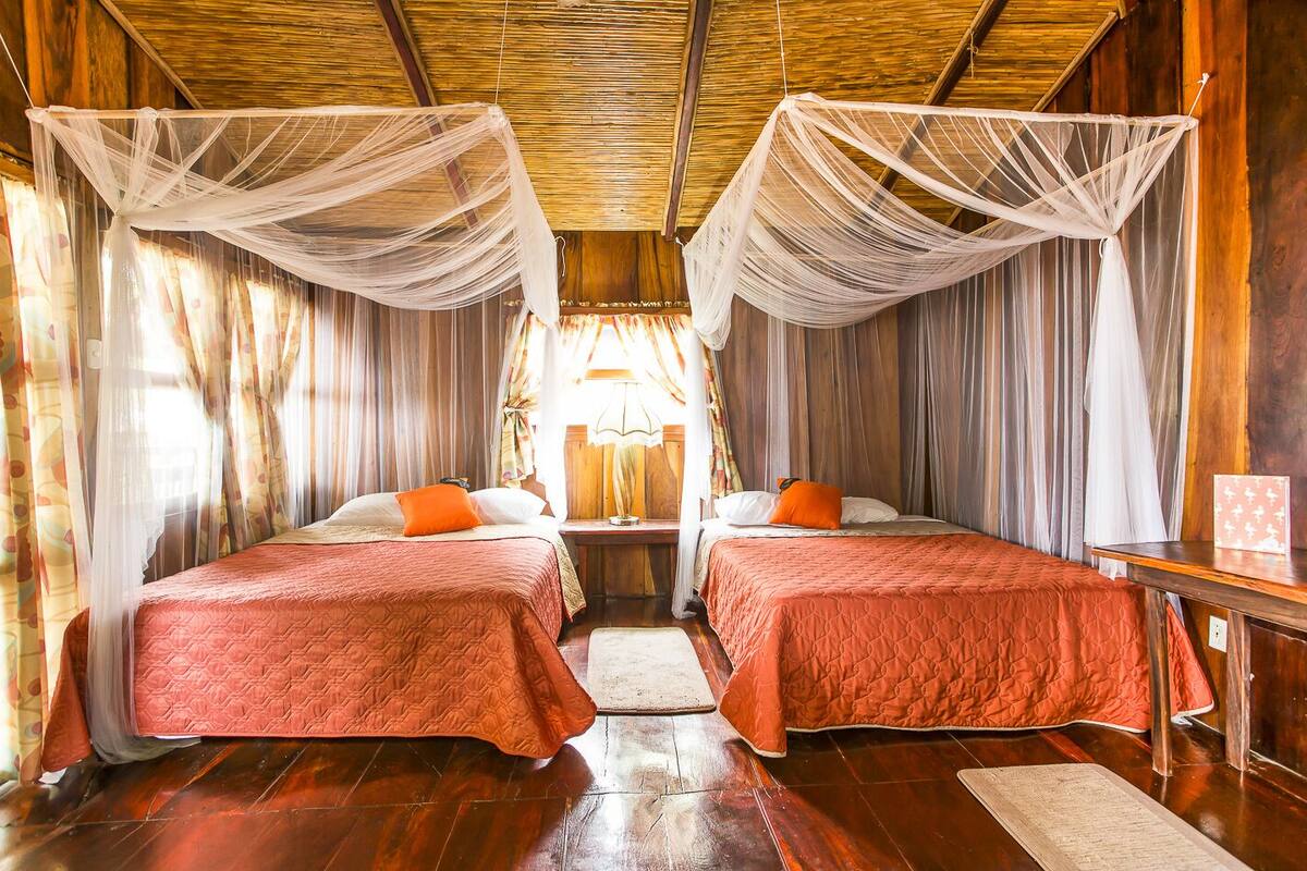 A room with two beds, each with a red quilted bedspread and orange pillows. There is a wooden floor and a small table with a lamp.