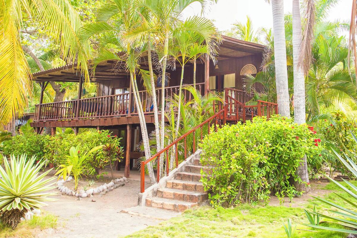 A wooden structure with a balcony and stairs leading to a covered area, surrounded by lush tropical vegetation.
