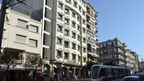 A multi-story hotel with a classic architectural style, featuring a prominent entrance with a sign, and a tram passing by on the adjacent street. - Assala (Oran)