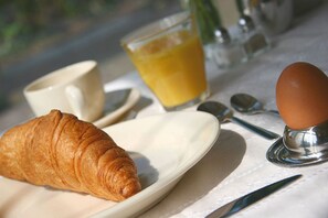 Disposição para o pequeno-almoço com um croissant, uma chávena, um copo de sumo de laranja e um ovo cozido sobre uma mesa.