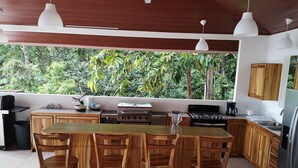 A modern kitchen with wooden cabinets, a central island with bar stools, a gas stove, a coffee maker, and a view of lush greenery. - Casa Odi-SEA - Portasol Vacation Rentals (Savegre)