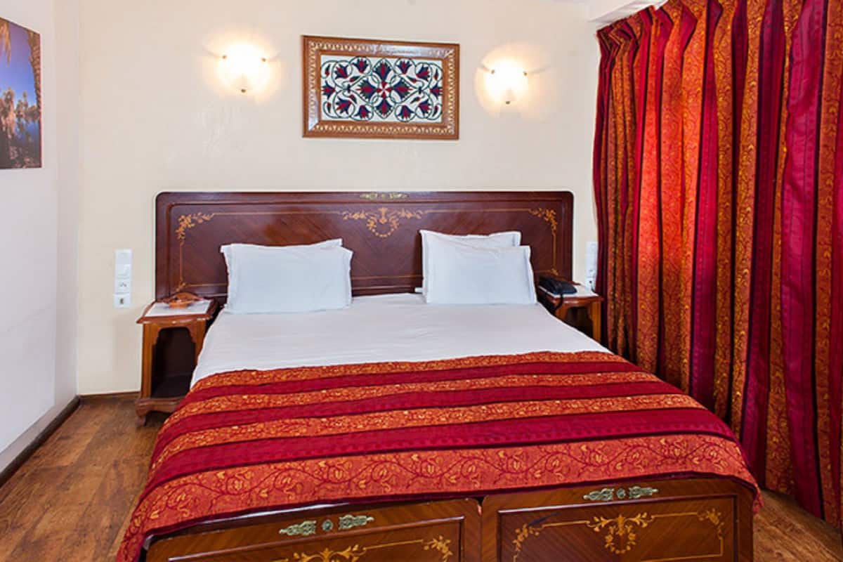 A hotel room with a wooden bed, two bedside tables, a nightstand, a patterned wall hanging, and a red curtain.