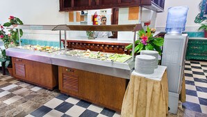 Zona de bufé con varias estaciones de comida, un dispensador de agua y una pila de platos sobre una mesa.