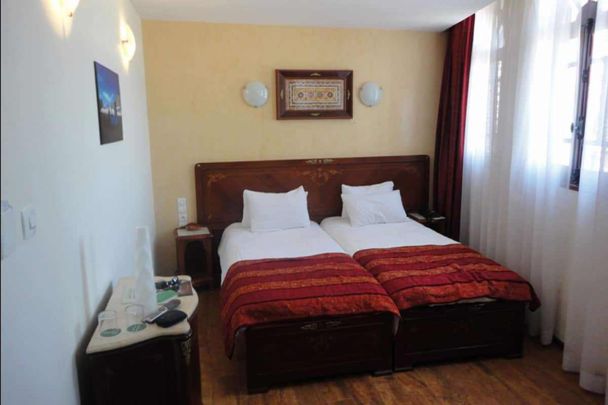 A hotel room with two beds, a nightstand, a small table with a phone and glasses, and a wall-mounted picture.