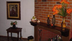 A room with a brick fireplace, a framed picture on the wall, a small table with a vase of flowers, and a shelf with candles and decorative items. - Hotel Villa Santo Domingo (Concepcion de Ataco)