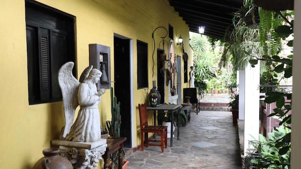 A covered outdoor area with a yellow wall, a statue, a table, chairs, and potted plants. - Hotel Villa Santo Domingo (Concepcion de Ataco)