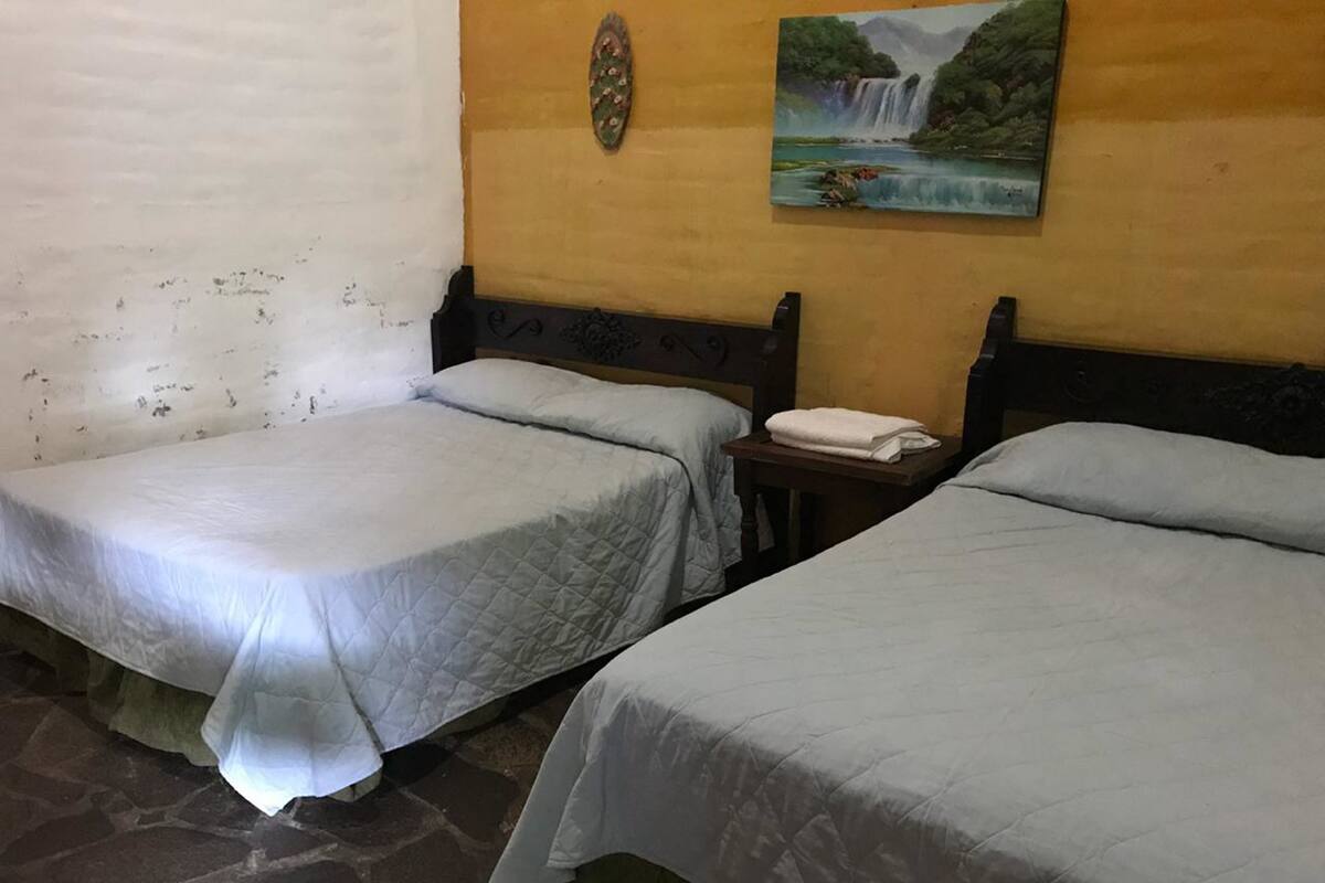 Two single beds with white linens, a wooden headboard, a small table with a folded towel, and a painting of a waterfall on the wall.