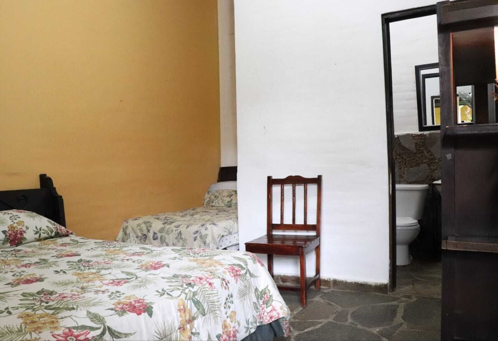 Una habitación con dos camas, una silla de madera y un baño visible a través de una puerta abierta.