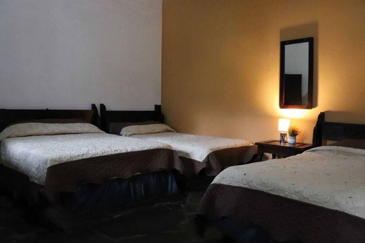 A hotel room with three single beds, a nightstand with a lamp, and a mirror on the wall.