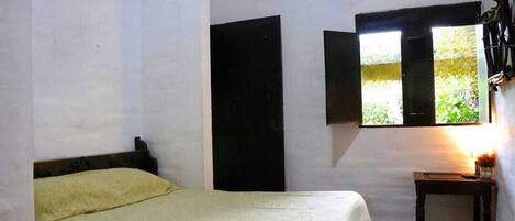Quarto com cama, janela com persianas abertas e pequena mesa com abajur.