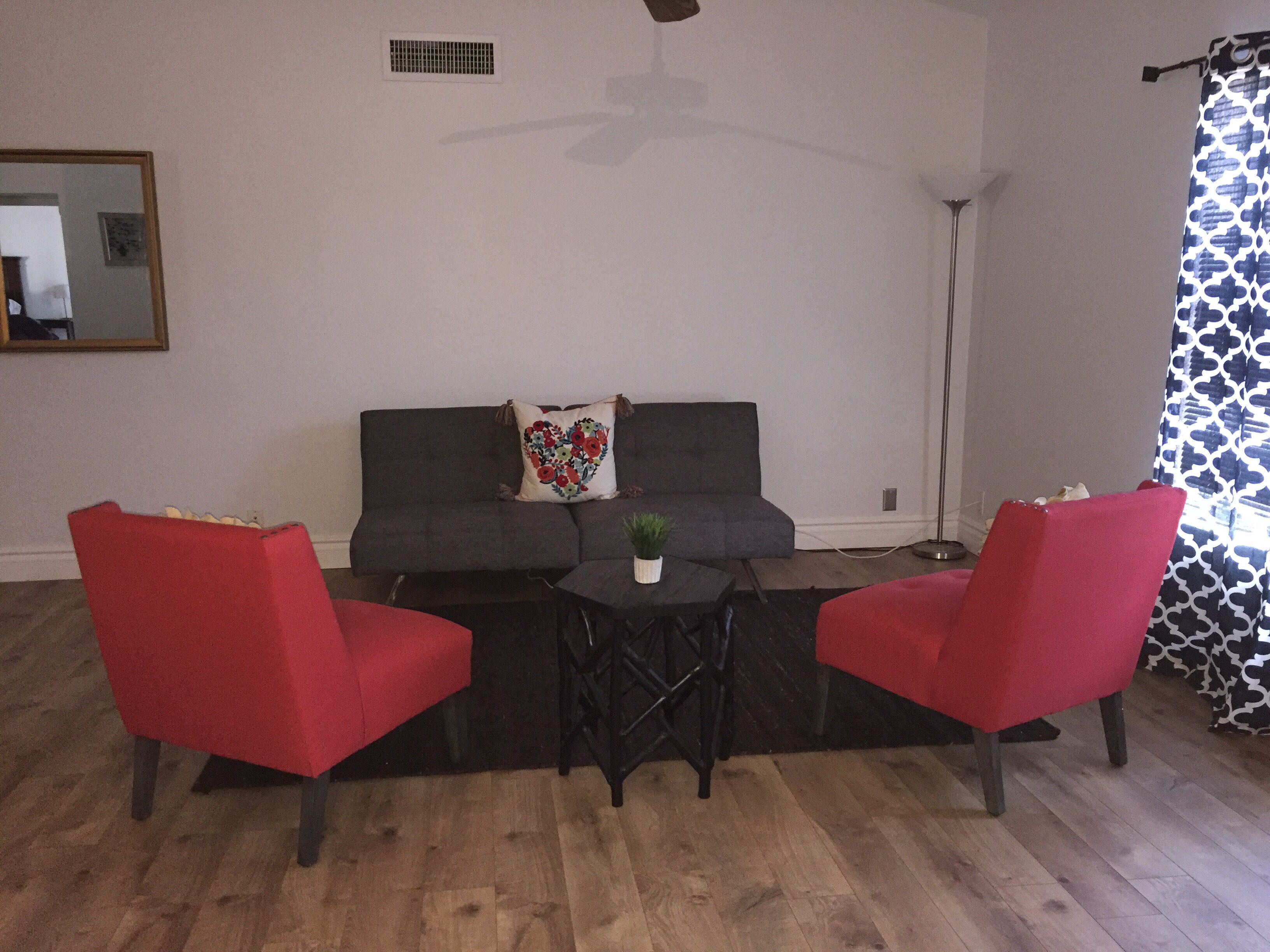 Een woonkamer met een grijze bank, twee rode stoelen, een kleine tafel met een plant en een plafondventilator.