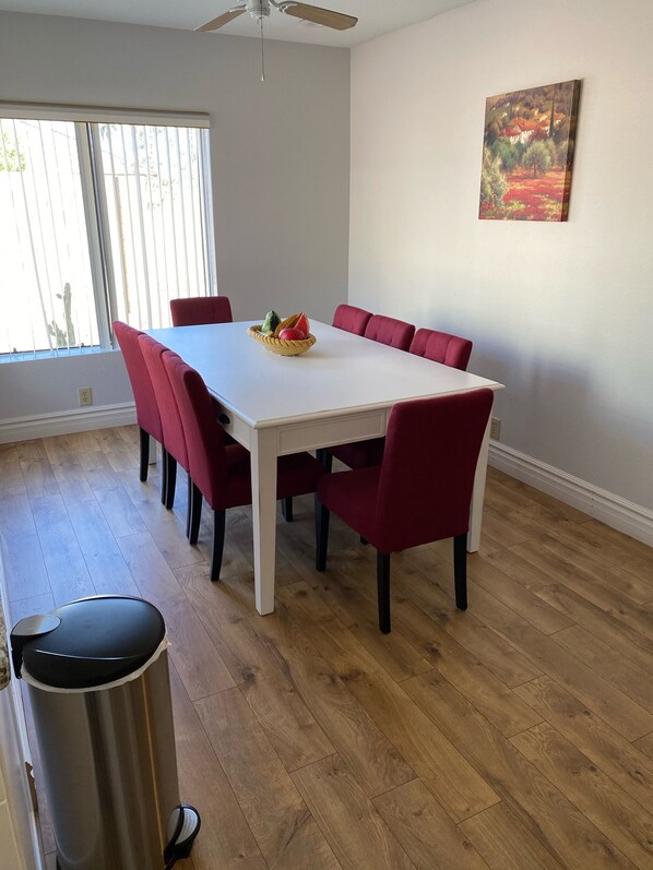 Un coin repas avec une table blanche, des chaises rouges, un panier de fruits et un tableau accroché au mur.