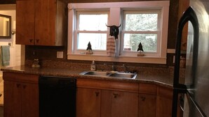 A kitchen with wooden cabinets, a stainless steel refrigerator, a double sink, and a window with a view of trees.