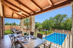 Un área de comedor al aire libre con vista a la piscina, pilares de piedra y una hamaca colgante.