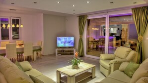 Una sala de estar moderna con una televisión de pantalla plana, asientos cómodos, un comedor y vistas a través de puertas de vidrio grandes.