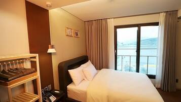 A hotel room with a bed, a wooden shelf with clothes, a nightstand with a phone, and a window with a view of a body of water.