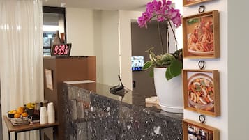 Ein Hotelrezeptionsbereich mit einer Marmor-Theke, einer Uhr, die 9:39 anzeigt, einer Blumenvase und gerahmten Bildern an der Wand.