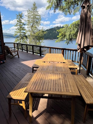Een houten terras met tafels en stoelen, met uitzicht op een meer met bomen en een bebost gebied op de achtergrond.