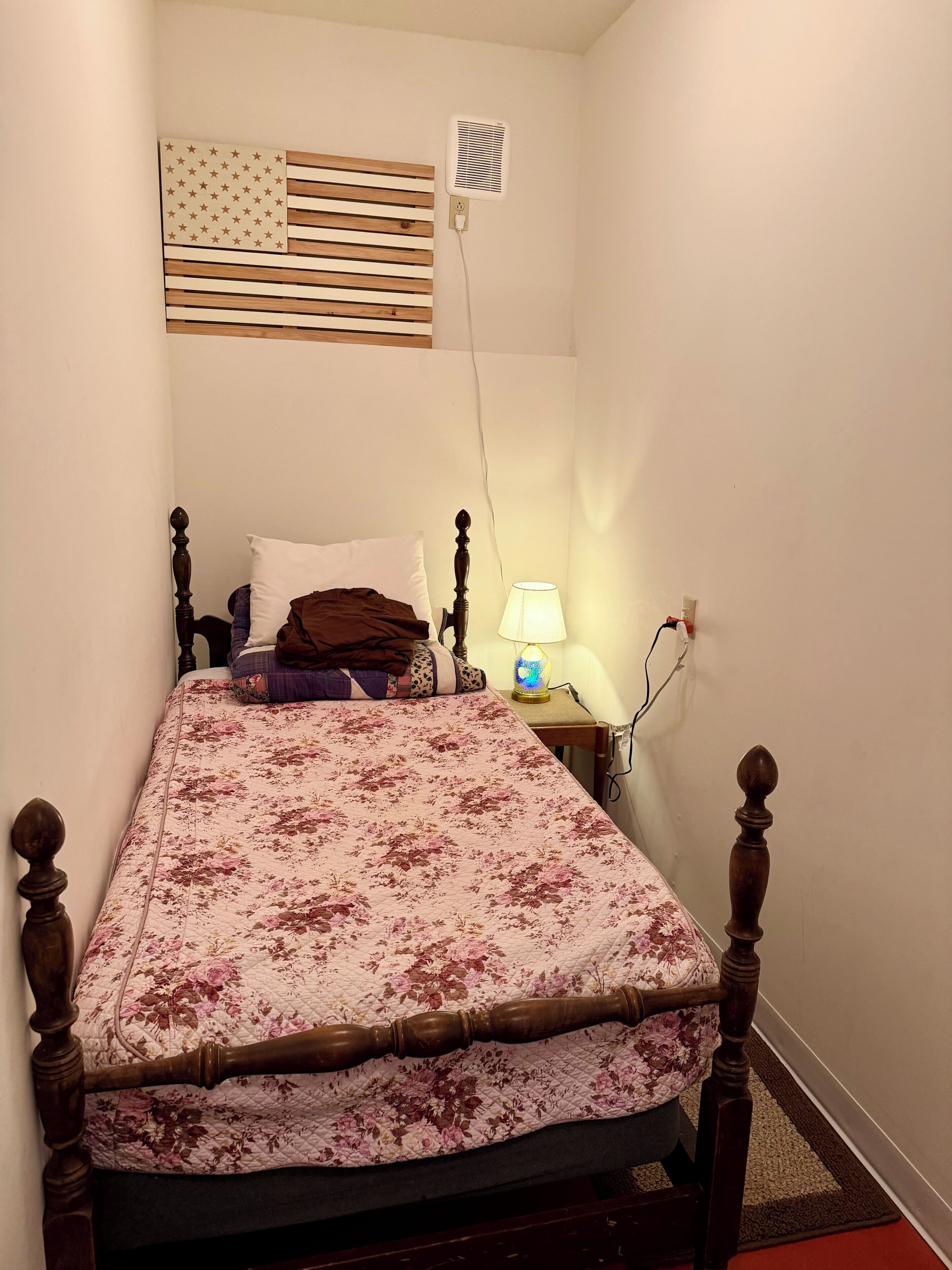 A single bed with a floral bedspread, a bedside lamp, a wall-mounted air vent, and a small table with a lamp.