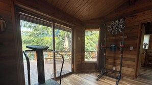 A wooden cabin interior with a treadmill, a clock, and a view of a deck and trees. - Spectacular 3-Story 7BR CDA Lakefront w-Dock on Secluded Powderhorn Bay (Harrison)