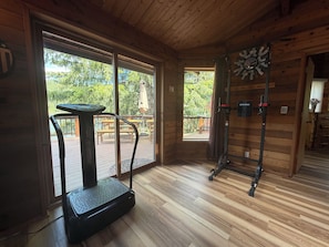 Interieur van een houten chalet met een loopband, een klok en uitzicht op een terras en bomen.