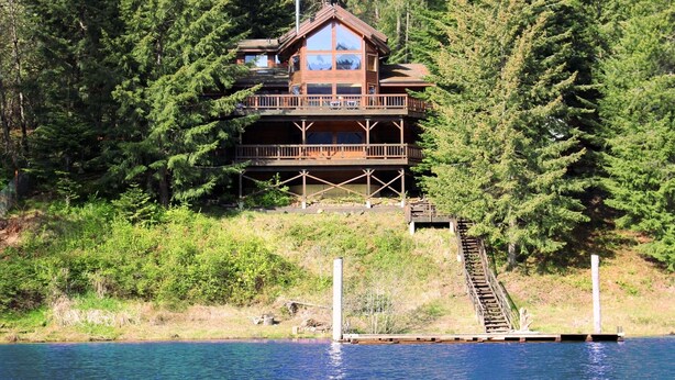 A multi-level cabin with a balcony, surrounded by dense forest, overlooking a body of water. - Spectacular 3-Story 7BR CDA Lakefront w-Dock on Secluded Powderhorn Bay (Harrison)