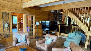 A log cabin interior with a wooden floor, two sofas with floral cushions, a wooden coffee table, a flat-screen TV, and a staircase. - Spectacular 3-Story 7BR CDA Lakefront w-Dock on Secluded Powderhorn Bay (Harrison)
