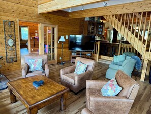 Een blokhutinterieur met een houten vloer, twee banken met bloemenkussens, een houten salontafel, een flatscreen televisie en een trap.