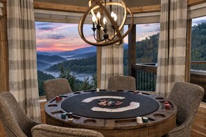 A round wooden table with chairs, a chandelier, and a view of a forested landscape through large windows.