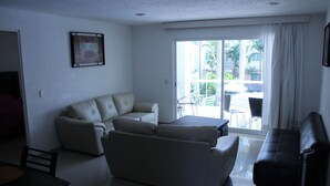 A living room with a sofa, a coffee table, and a TV. There is a sliding glass door leading to an outdoor area with chairs and a table. - Nice Apartment, perfect for families!  (Diamante Lakes)