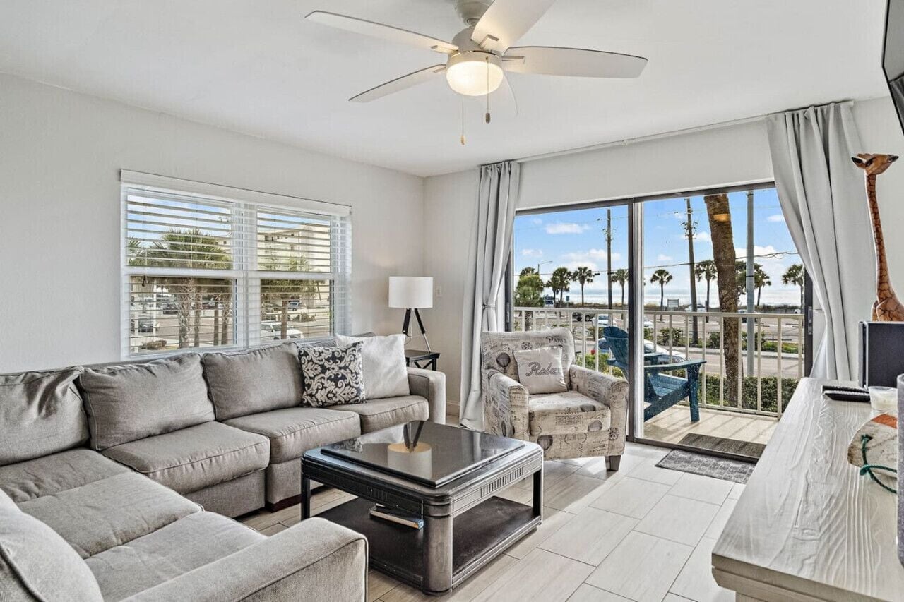 A modern living room with a sofa, coffee table, and armchair, large windows with blinds, a ceiling fan, and a view of palm trees and a beach.