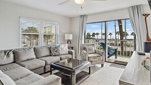 Un salón moderno con sofá, mesa de centro y sillón, ventanas amplias con persianas, un ventilador de techo y vistas a palmeras y a la playa.
