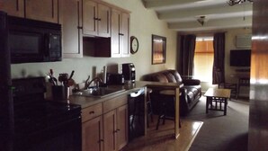 A kitchen with wooden cabinets, a microwave, and a coffee maker. There is a dining area with a table and chairs, and a living room with a couch and a TV. - Windjammer Lodge (Ogdensburg)
