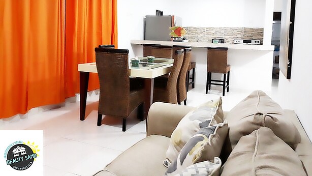 Een moderne woonkamer met een bank, eettafel en stoelen. Er is een keuken met een aanrecht en apparatuur. Oranje gordijnen zijn links te zien.