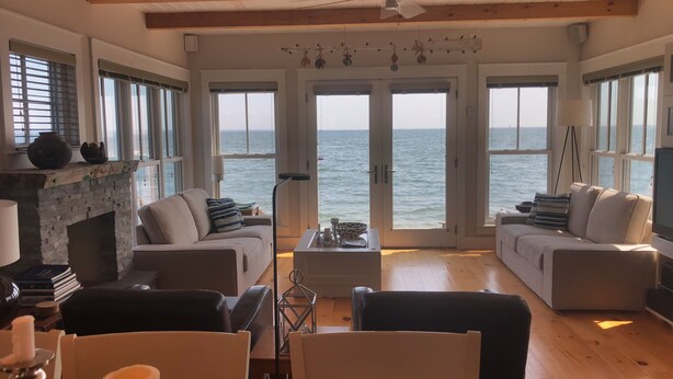 A spacious living room with a fireplace, two sofas, a coffee table, and a dining area with chairs. Large windows offer a view of the sea. - Living on the edge! Breathtaking waterfront living at it’s best!  (Provincetown)
