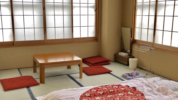 A traditional Japanese room with tatami flooring, a low wooden table, red cushions, a bed with a red floral bedspread, and a sliding door with a frosted glass panel. - Resort Inn Fujihashi (Fujikawaguchiko)