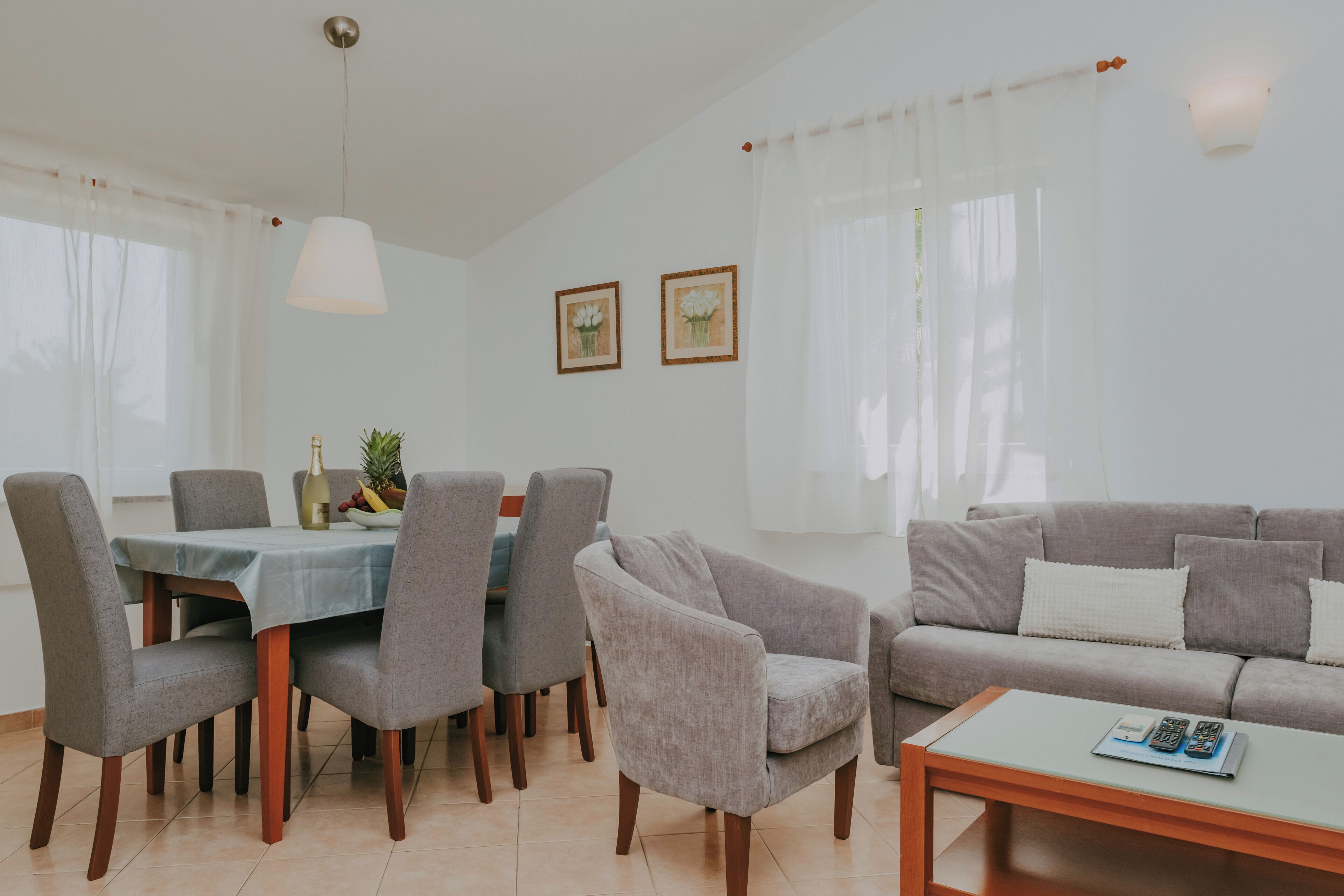 Ein Essbereich mit einem Tisch, gedeckt für vier Personen, umgeben von grauen Stühlen. Ein graues Sofa und ein Couchtisch mit einer Zeitschrift und Fernbedienungen stehen daneben.