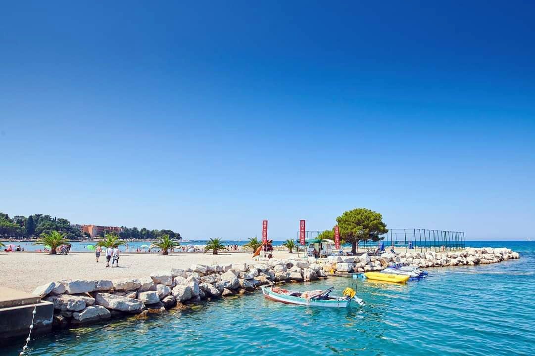 A coastal beach with clear blue water, rocky breakwaters, and small boats.