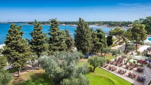 A coastal hotel with outdoor seating overlooking the sea, surrounded by trees and a clear sky. - Villa DANEX STANDARD **** (Umag)