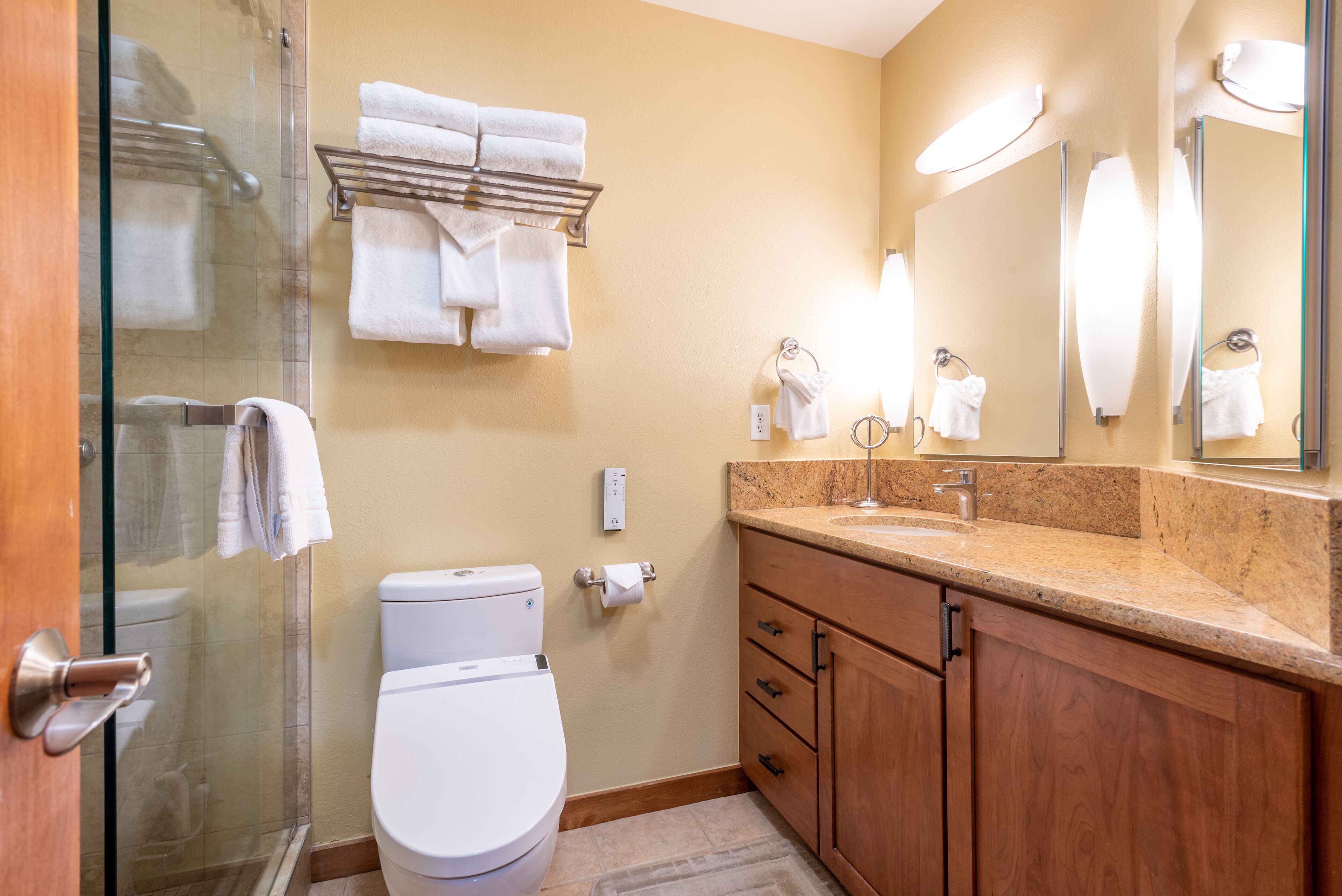 A bathroom with a glass shower enclosure, a white toilet, a wooden vanity with a granite countertop, and a mirror with lights.
