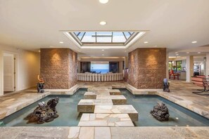 Un vaste espace intérieur abritant une piscine, agrémenté d’un élément aquatique central, de murs en pierre et d’une verrière.