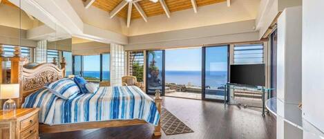 A bedroom with a large bed, wooden flooring, a ceiling fan, a TV, and a balcony with a view of the ocean.