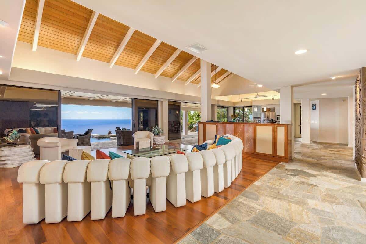 Uma sala ampla com um sofá branco curvo, uma mesa de centro de vidro e teto de madeira.