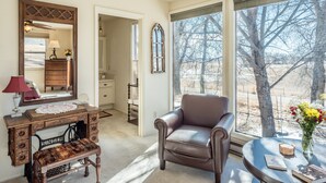 Ein Zimmer mit großem Fenster, braunem Ledersessel, Holzschrank, Spiegel und Blumen in einer Vase.