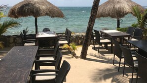 A beachside dining area with tables and chairs, thatched umbrellas, and a view of the ocean.